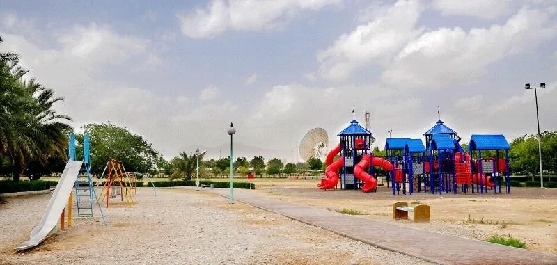 Children's playground with slide and swings on gravel, palm trees and the Muscat satellite station dishes in the background at Al Amerat Public Park