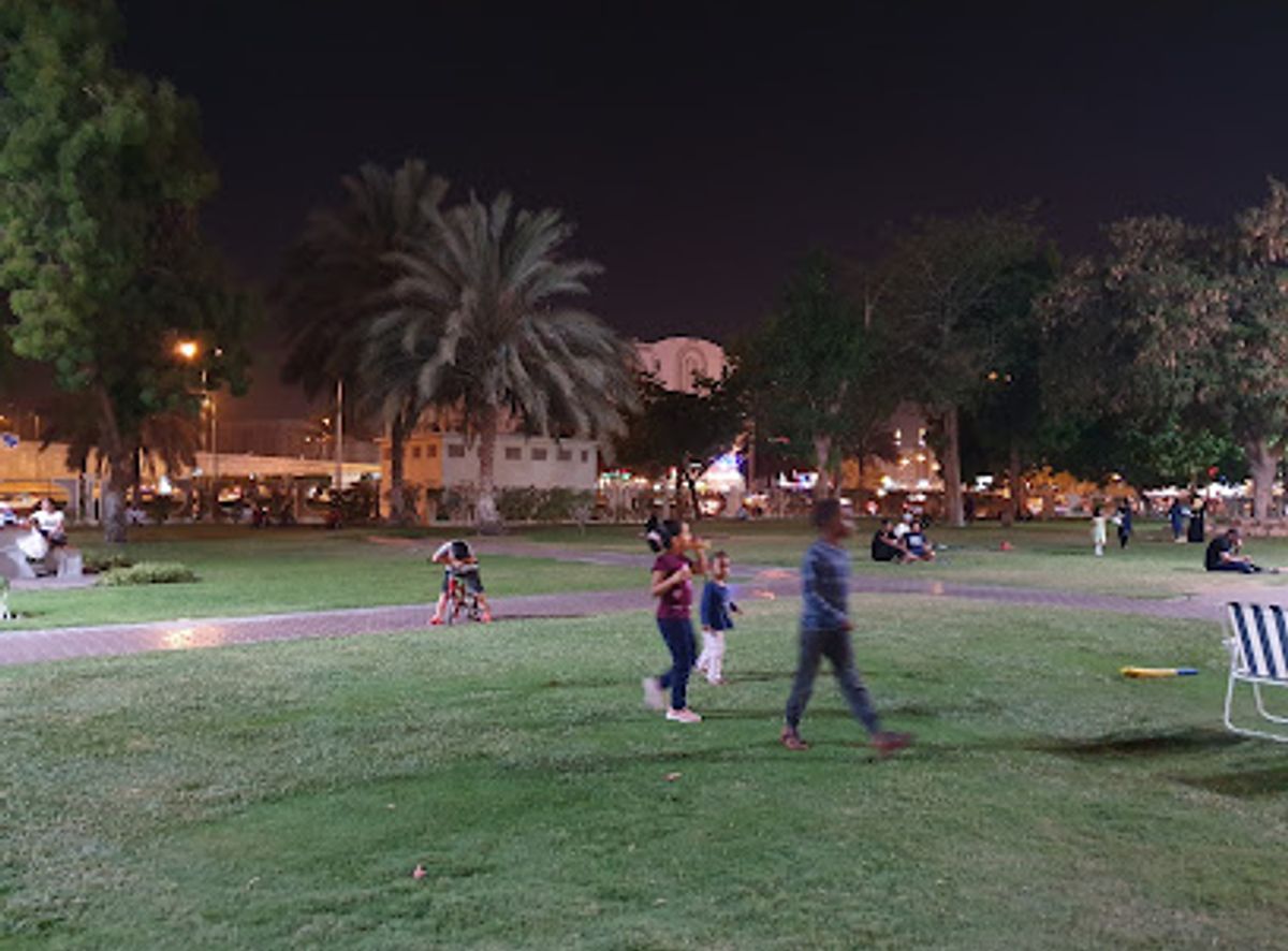 Families with children on the lawn at Al Khuwair Central Park at night, lit paths and palm trees in the background