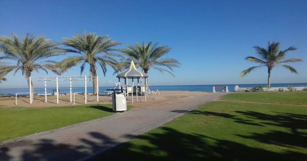 Outdoor exercise equipment, a shaded gazebo and palm trees leading onto the sand at Azaiba Beach Park
