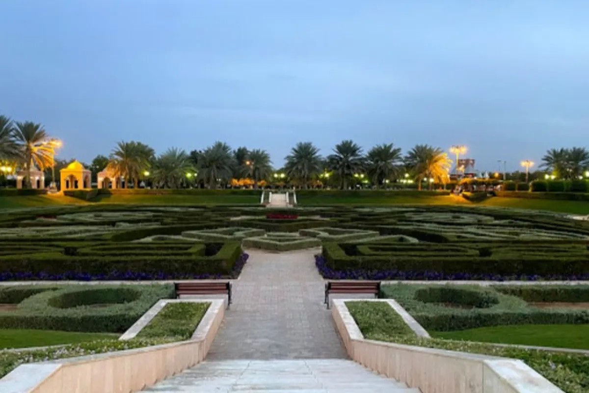 Formal clipped-hedge garden with a paved path, benches and gazebos lit at dusk in Al Sahwa Public Garden, Seeb