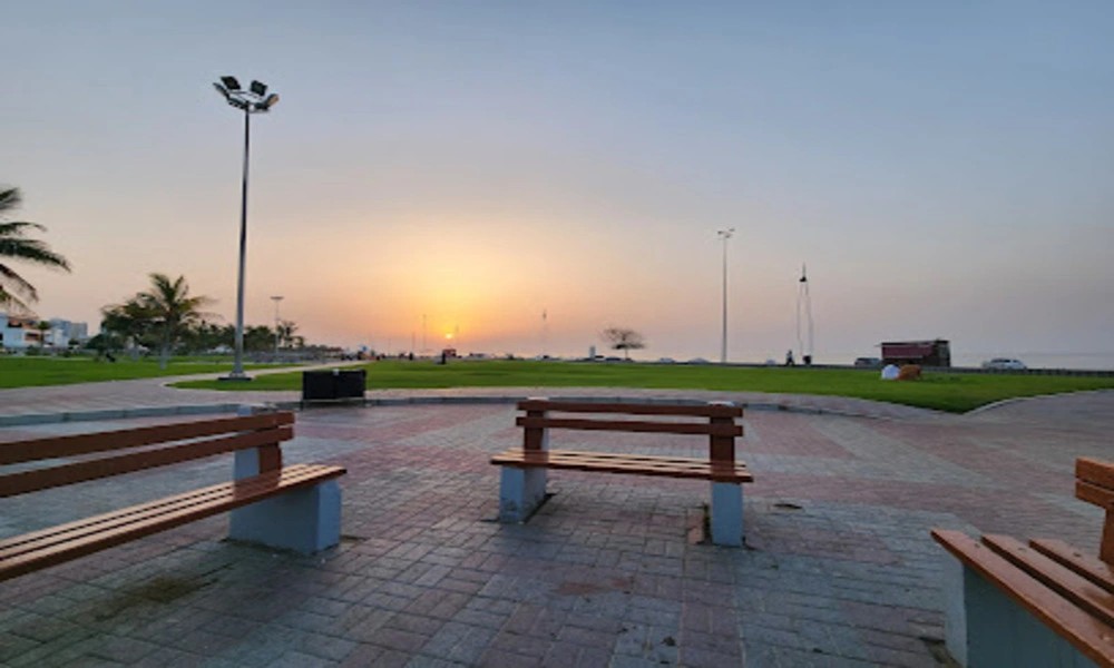 Wooden benches on a paved terrace overlooking the lawn and beach at Ghubrah Beach Park at sunset