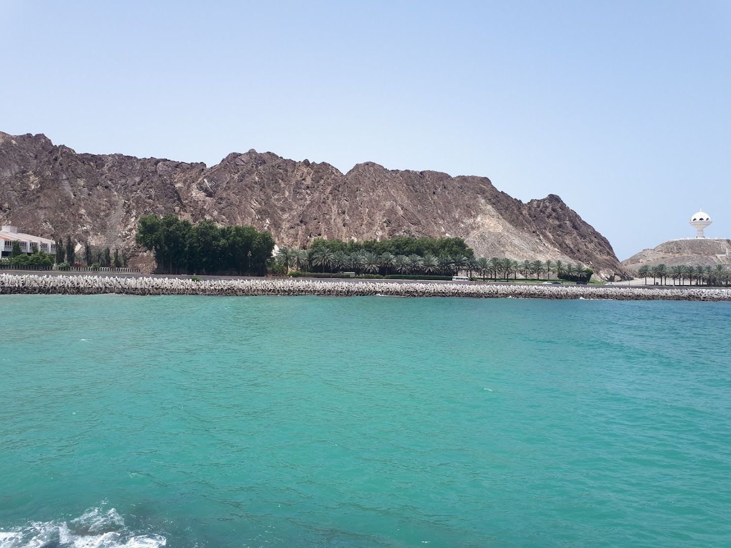 View from the water of the palm-lined seawall path and lawn at Kalbuh Park on the Muttrah Corniche, with the Riyam headland in the distance
