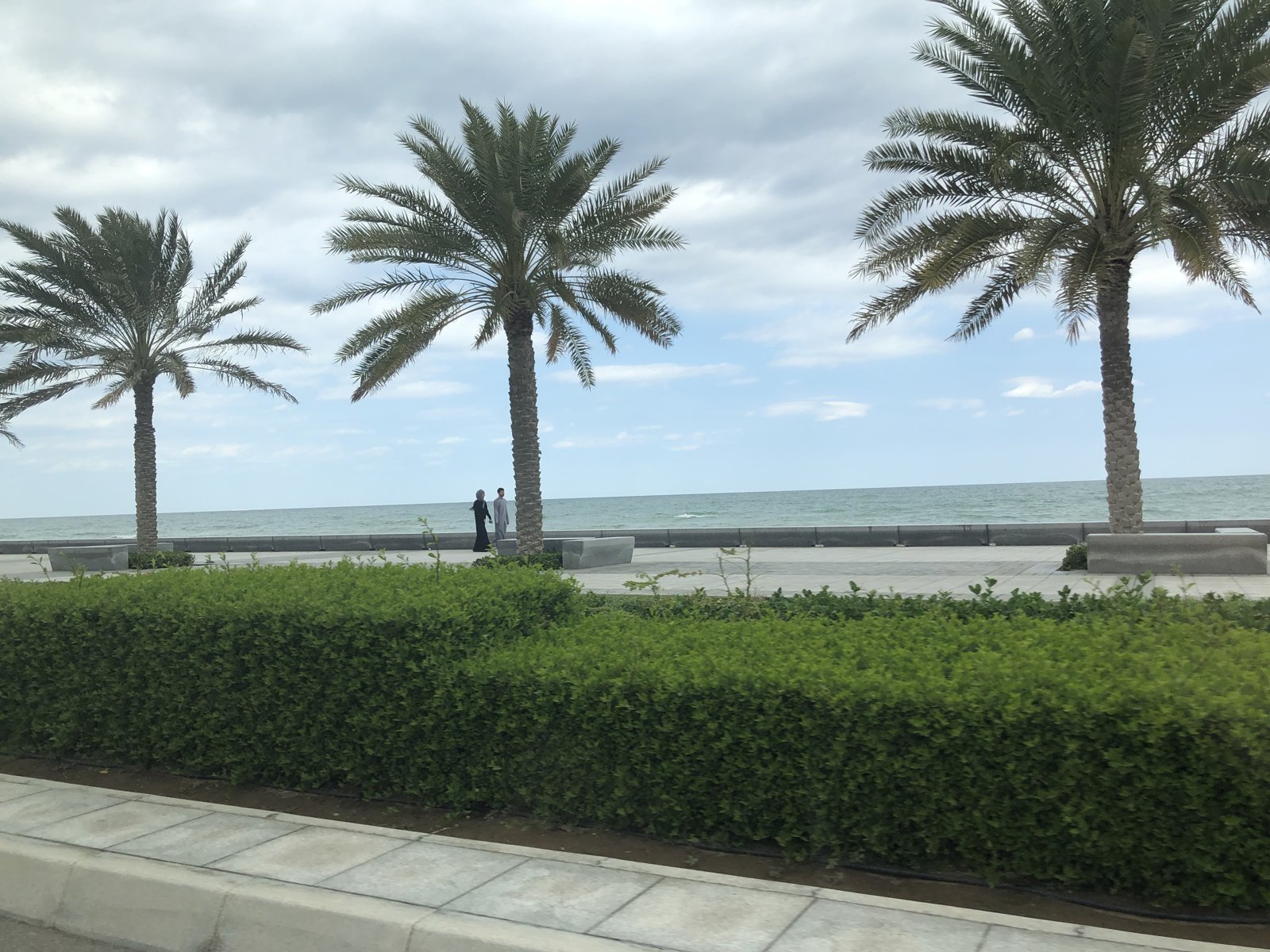 Palm-lined seafront promenade in Seeb with a couple walking beside the sea wall and the Arabian Sea beyond