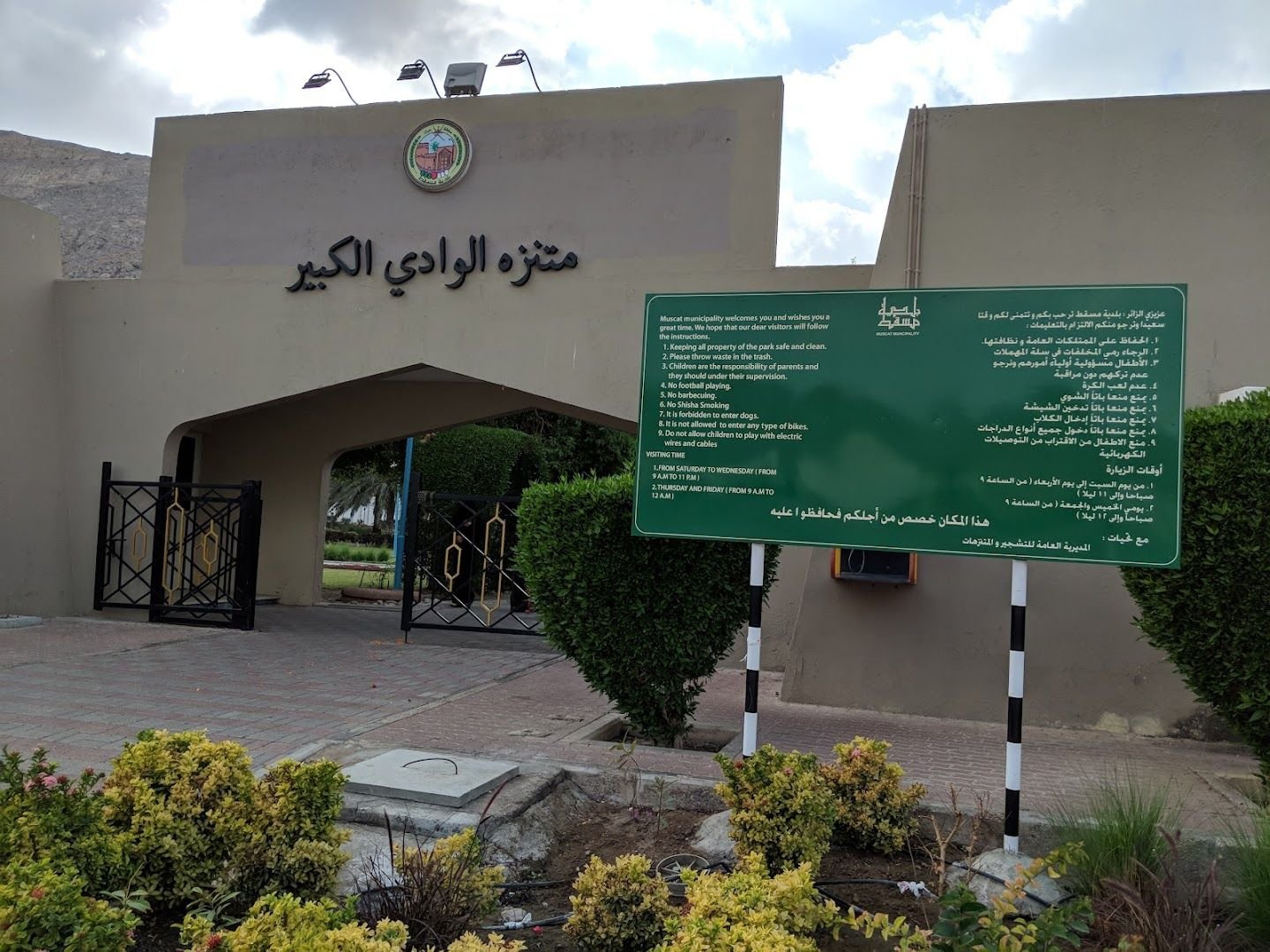 Park entrance gate with the official Arabic sign reading Muntazah Al Wadi Al Kabir and the posted Muscat Municipality visiting hours and rules
